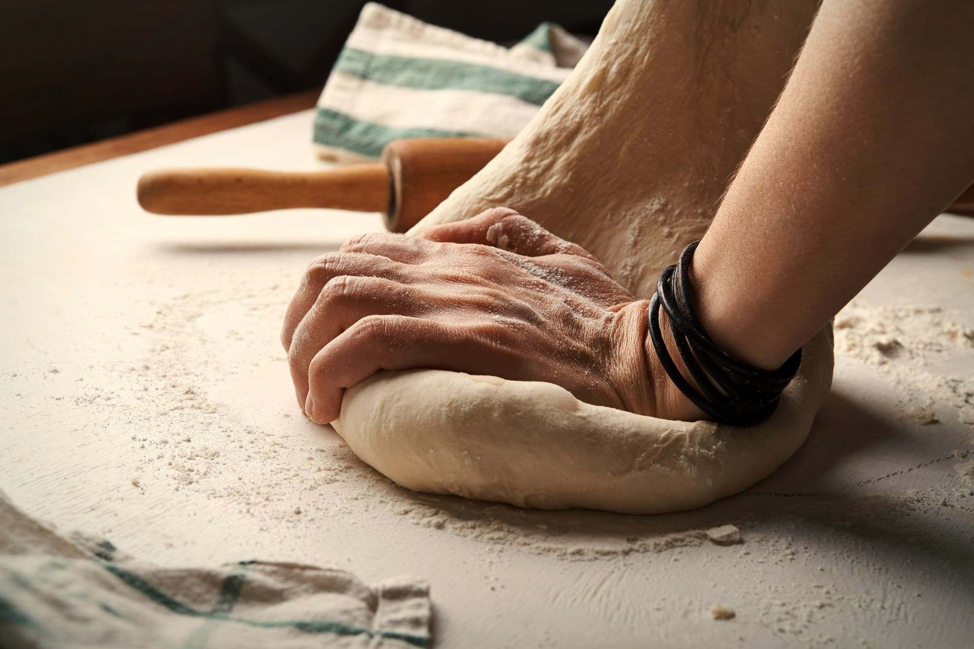 A baker's hand pressing a round of dough on a floured bench beside a rolling pin.