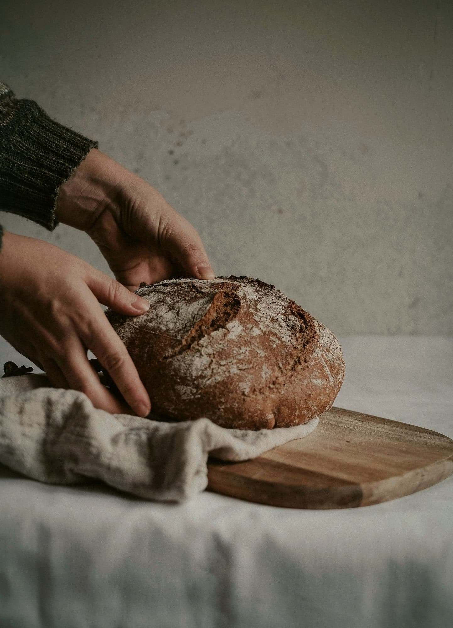Hands steadying a dark, floured sourdough loaf on a linen cloth.