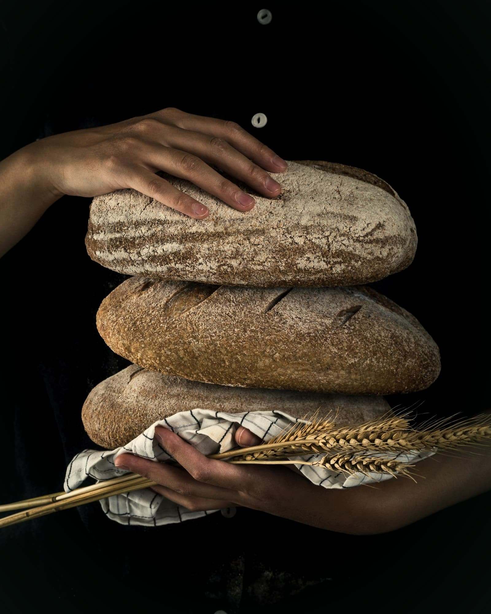 A stack of three seeded rye loaves held in a baker's hands, with ears of wheat.