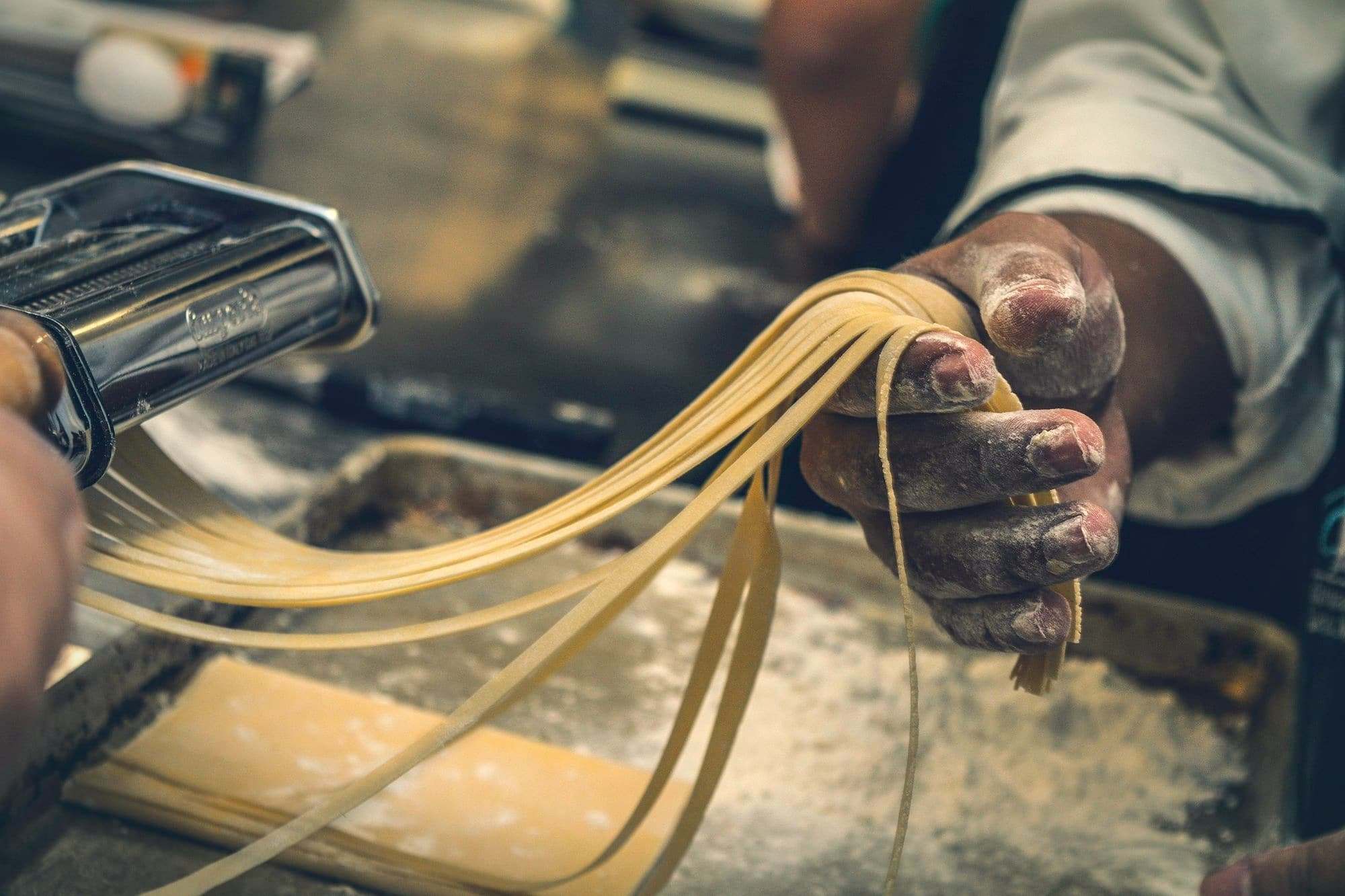 A cook's floury hand drawing ribbons of fresh pasta from a rolling machine.