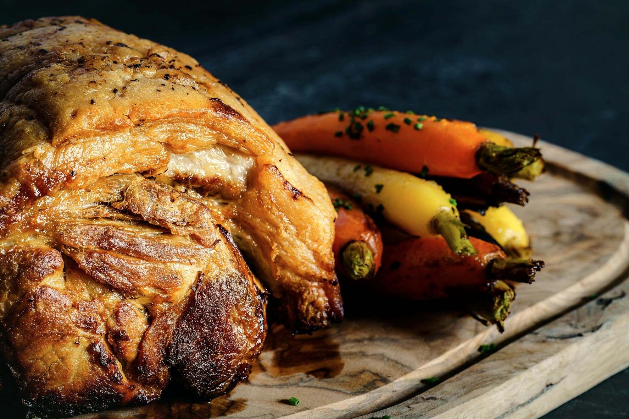 A roast joint with crisp crackling and glazed carrots on a wooden board.