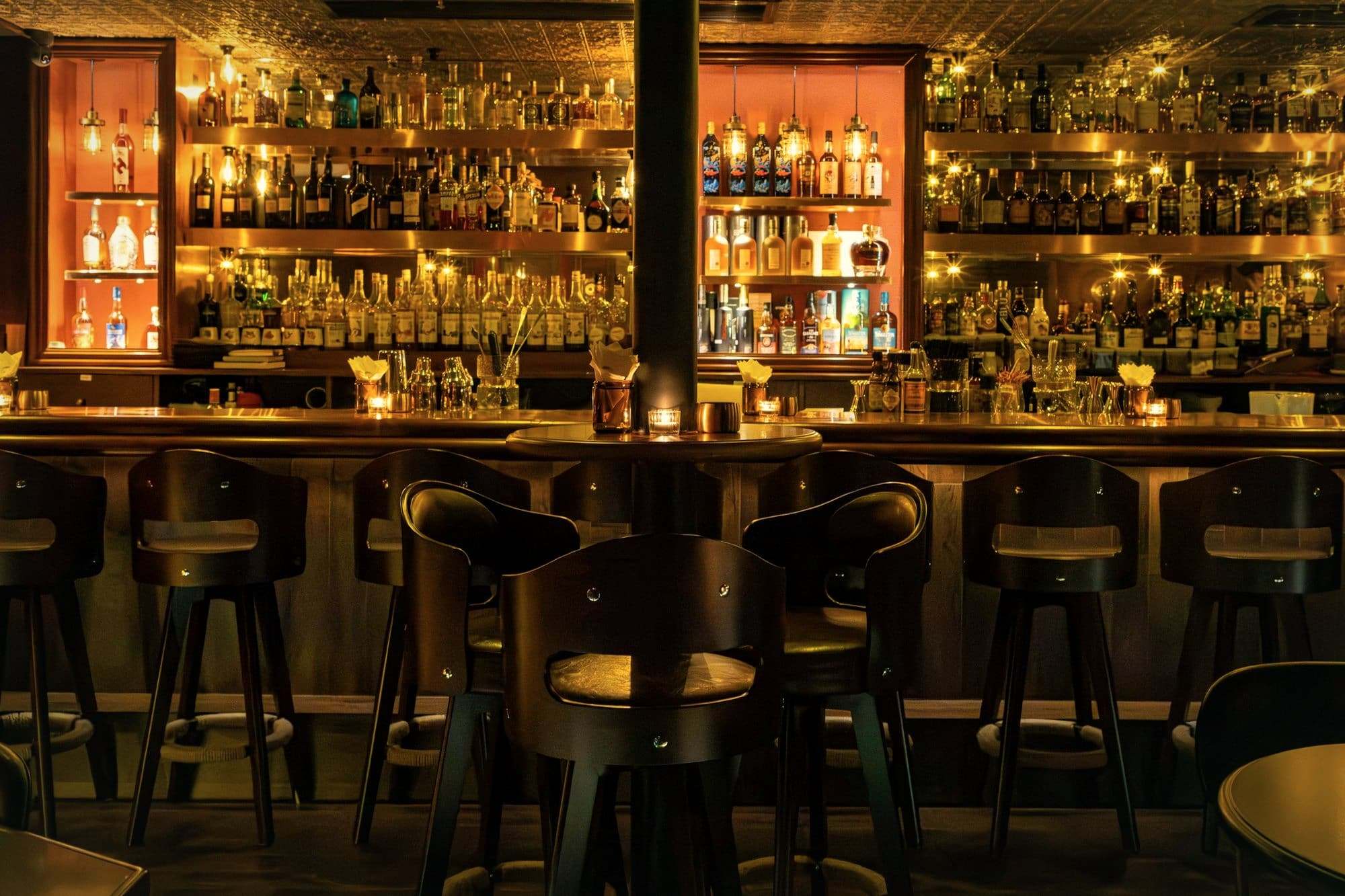 Bar stools and shelves of bottles lit amber at night.