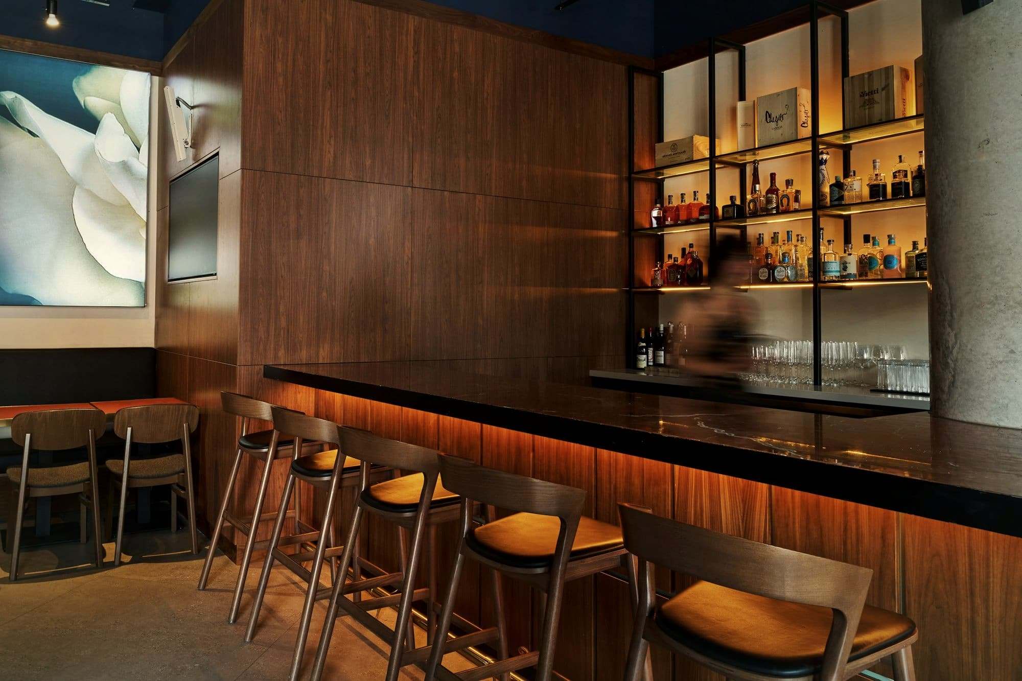 A row of stools along a dark wooden bar with lit shelves behind it.